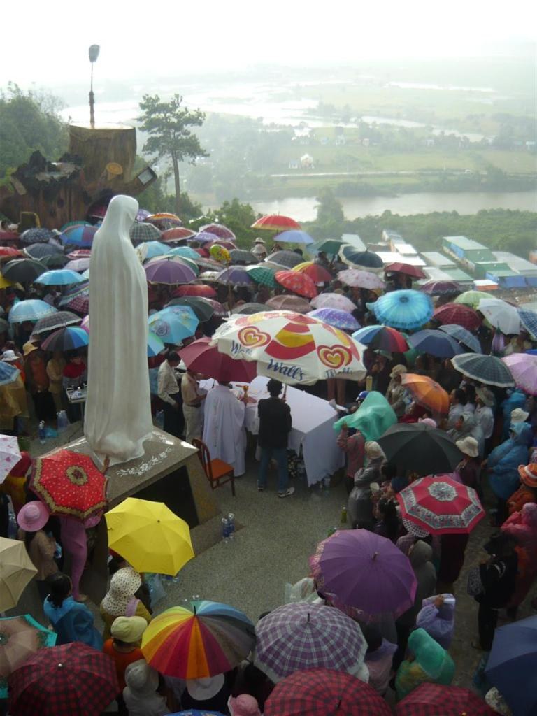 Đông đảo khách hành hương che ô quanh linh đài tượng Đức Mẹ Tà Pao trong một buổi cử hành ngoài trời, nhìn xuống thung lũng phía dưới núi.